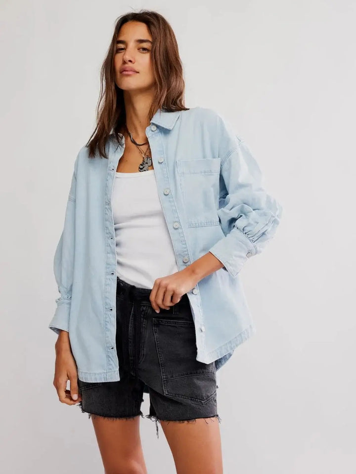 A model showcases the relaxed, oversized fit of the unbuttoned Free People Ashley Denim Shirt - Classic Blue Wash styled over a white tank top and black shorts | Twentyseven Toronto