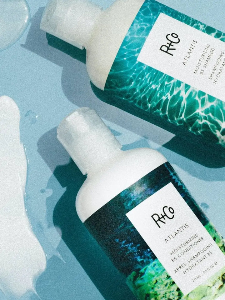 A bottle of R+Co ATLANTIS B5 Moisturizing Shampoo is pictured from above alongside its matching conditioner and a smear of white product on a light blue surface | Twentyseven Toronto