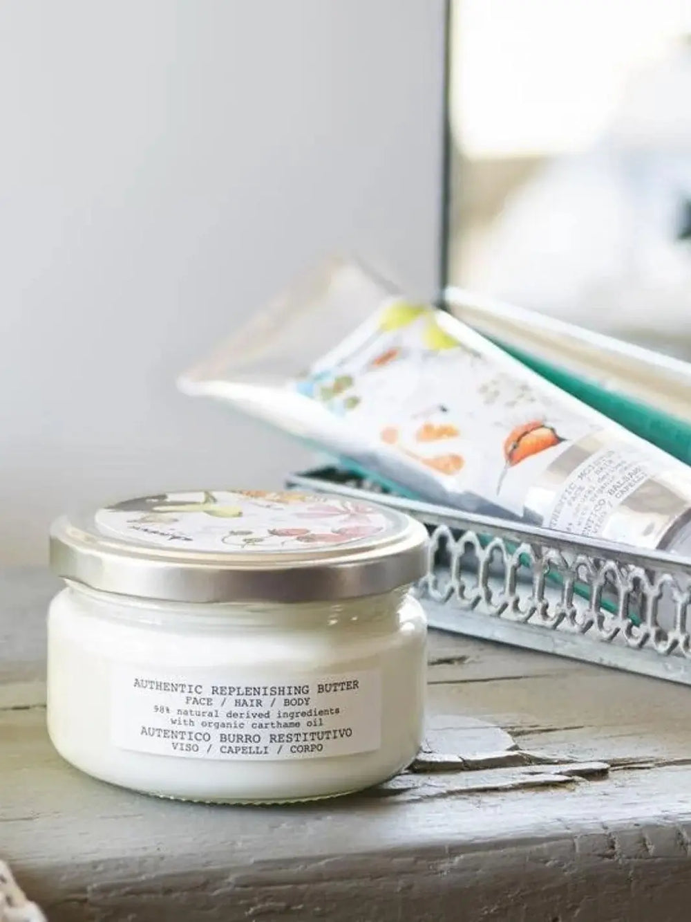 A clear glass jar of Davines Authentic Replenishing Butter for Face, Hair & Body sits on a rustic wooden surface with other beauty products blurred in a silver tray behind it | Twentyseven Toronto