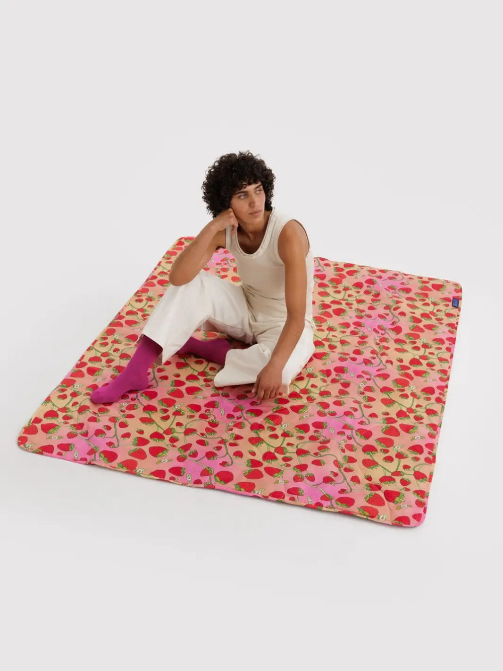 A person laying on the Baggu Puffy Picnic Blanket in Strawberry Blossom print featuring a pink and peach gradient background layered with an all-over red strawberry and green vine print | Twentyseven Toronto 