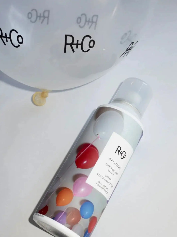 An aerosol can of R+Co BALLOON Dry Volume Spray lies on a white surface next to a white balloon printed with the R+Co logo | Twentyseven Toronto