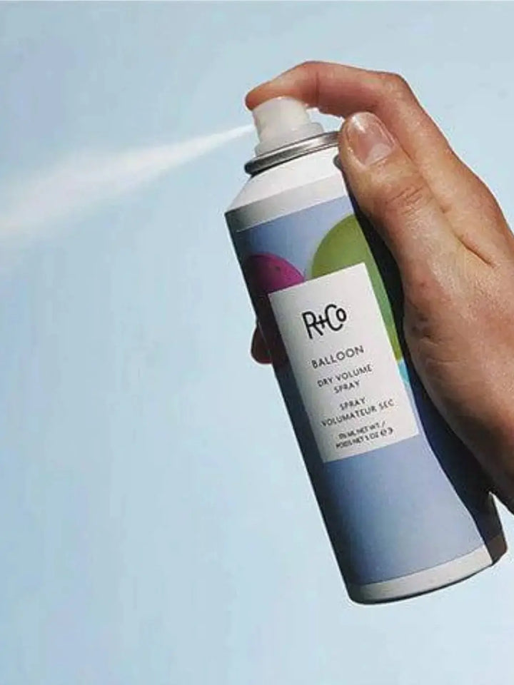 A hand presses down on the nozzle of an R+Co BALLOON Dry Volume Spray can, releasing a fine white mist against a solid blue background | Twentyseven Toronto