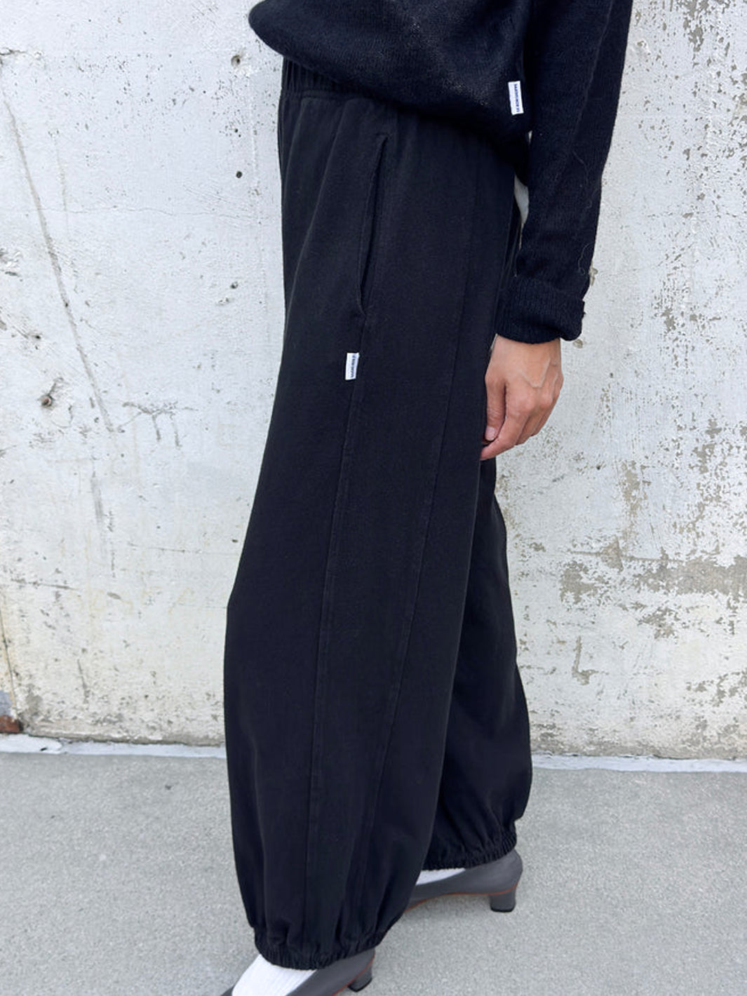 Side view of model wearing black Balloon Pants with a relaxed, voluminous silhouette, side pockets and elasticated cuffs, styled with a black sweater and grey heels | Twentyseven Toronto