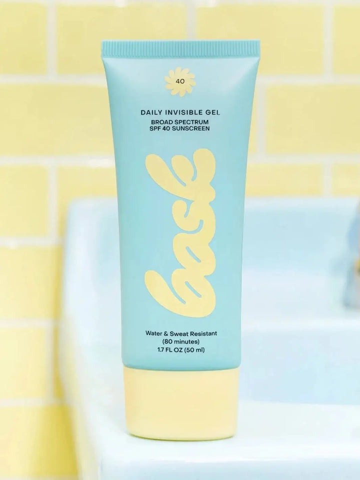 A light blue tube of Bask Suncare Daily Invisible Gel SPF 40 Sunscreen stands upright against a background of soft yellow tiles and a blue bathroom sink | Twentyseven Toronto