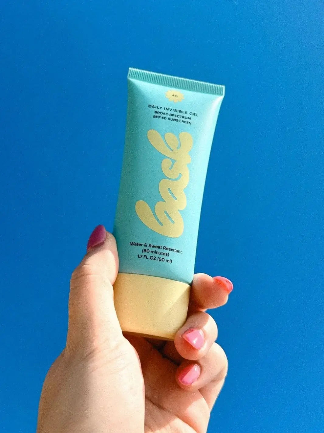 A hand with pink manicured nails holds a light blue tube of Bask Suncare Daily Invisible Gel SPF 40 Sunscreen up against a solid blue background | Twentyseven TOronto