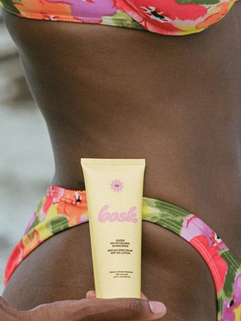 A person in a colorful floral bikini holds a yellow tube of Bask Suncare Bask SPF 50 Lotion Sunscreen against their hip | Twentyseven Toronto