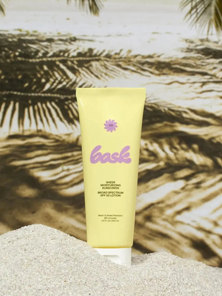A yellow tube of Bask Suncare Bask SPF 50 Lotion Sunscreen stands upright in white sand against a beach backdrop and palm trees | Twentyseven Toronto