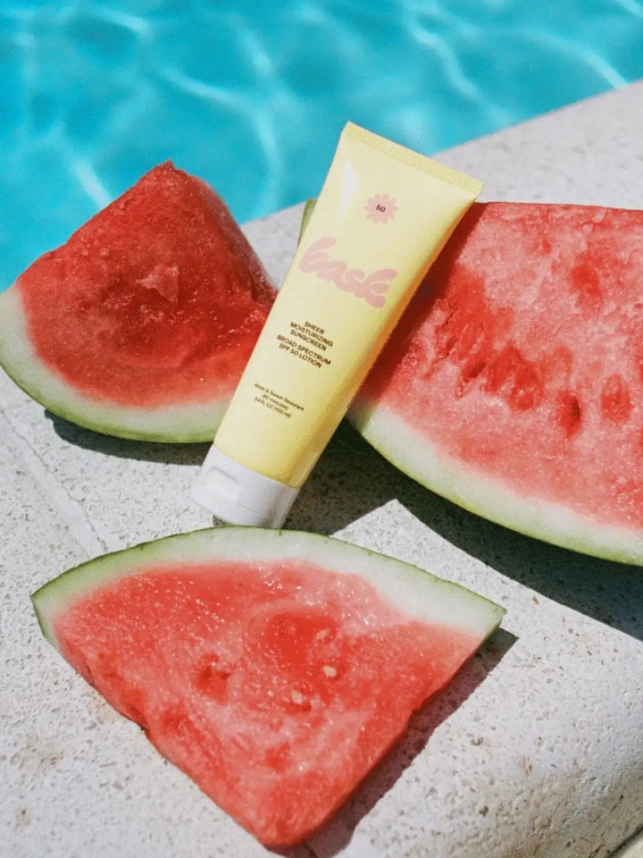 A yellow tube of Bask Suncare Bask SPF 50 Lotion Sunscreen rests on a concrete surface next to fresh slices of watermelon by the edge of a blue swimming pool | Twentyseven Toronto