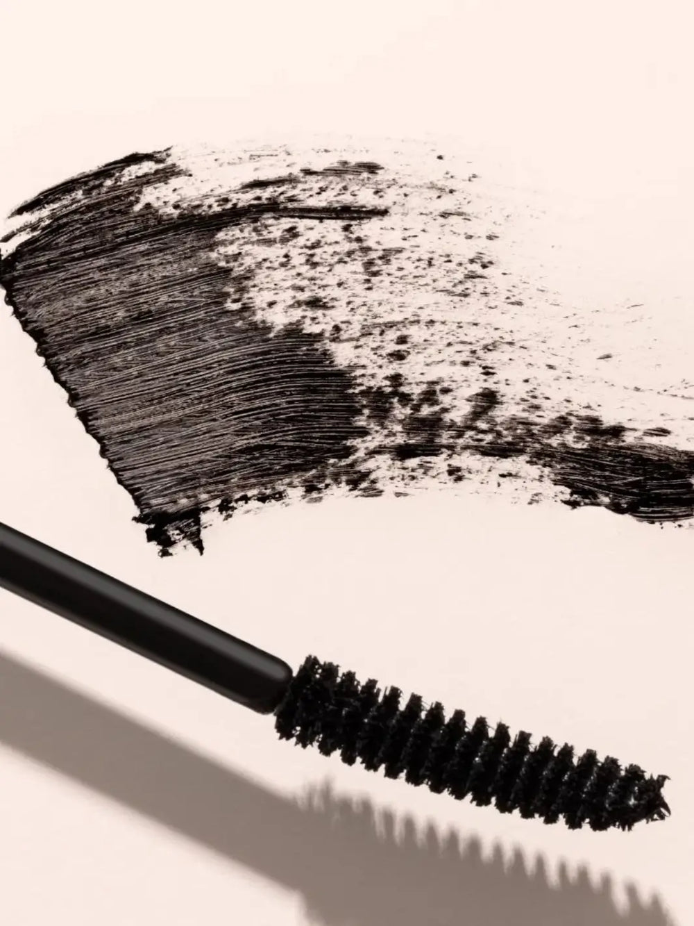 Close-up of the applicator wand for Beauty from Bees Mascara in Black resting beside a textured black pigment swatch on a light pink background | Twentyseven