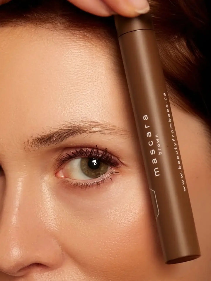 A woman holding a tube of Beauty from Bees Mascara in Black next to her face, highlighting her applied eye makeup | Twentyseven