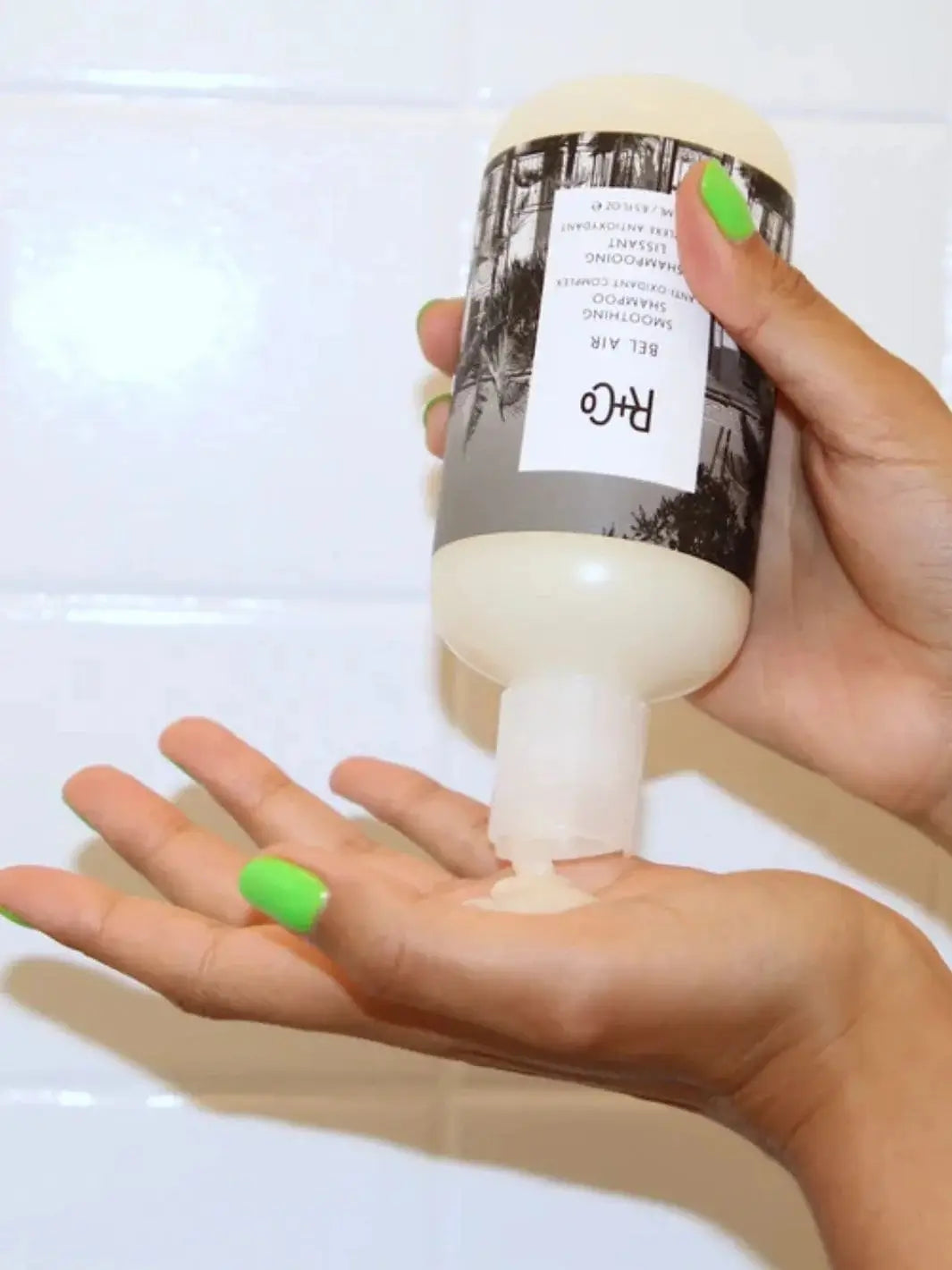 A person with bright green nail polish squeezes product from a bottle of R+Co BEL AIR Smoothing + Anti-Oxidant Complex Shampoo directly into their open hand | Twentyseven Toronto