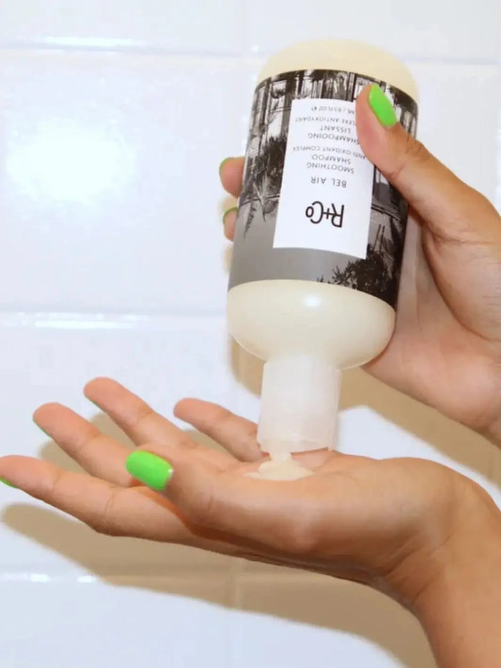 A person with bright green nail polish squeezes product from a bottle of R+Co BEL AIR Smoothing + Anti-Oxidant Complex Shampoo directly into their open hand | Twentyseven Toronto