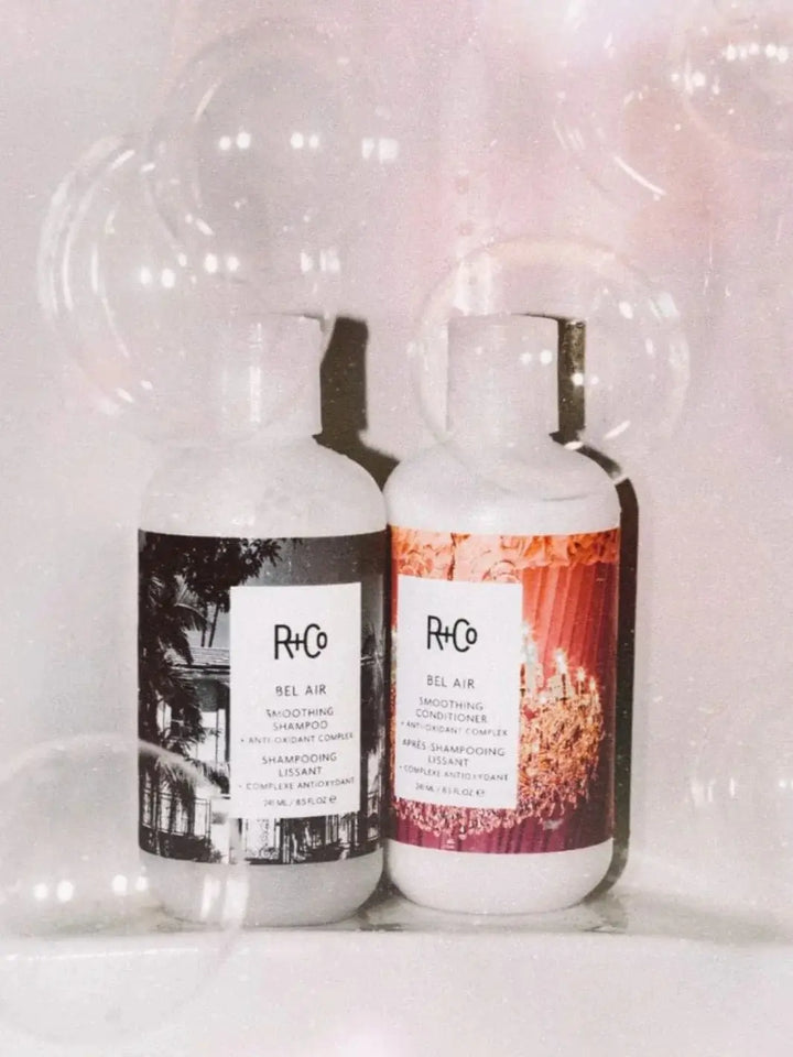 Two bottles of R+Co BEL AIR Smoothing + Anti-Oxidant Complex Shampoo and Conditioner stands on the edge of the tub surrounded by floating clear bubbles | Twentyseven Toronto