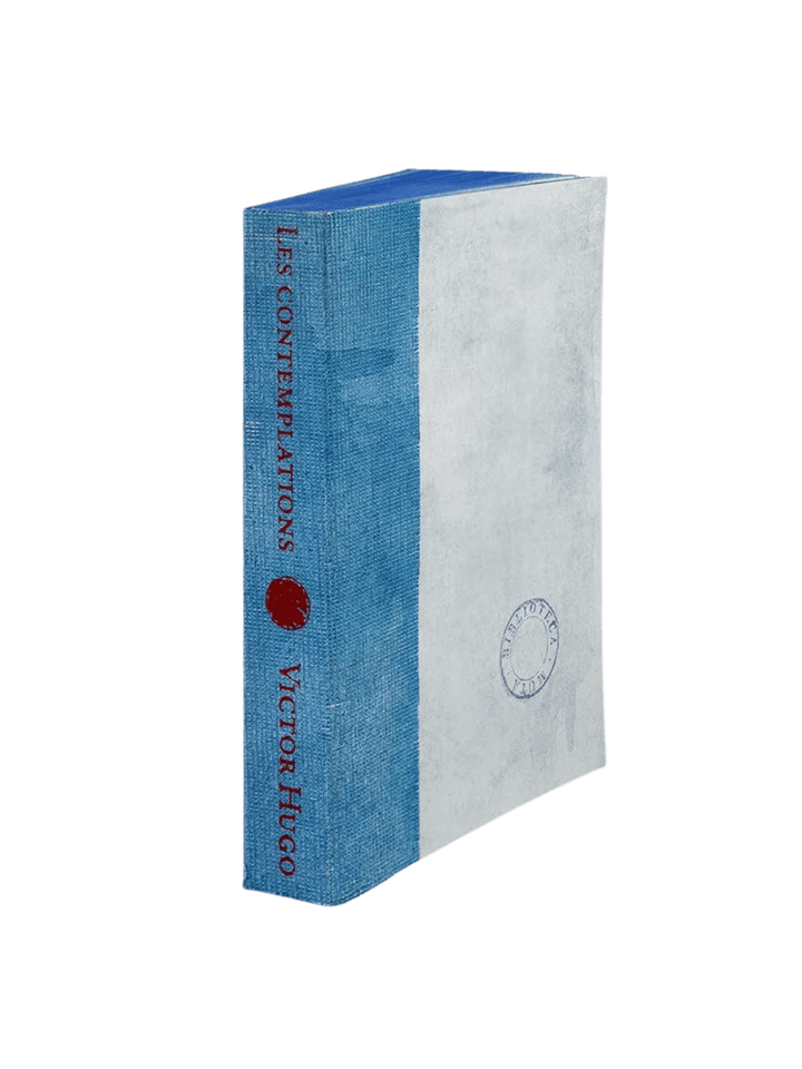 A notebook from the Les Contemplations from the Slow Design Biblioteca Muta collection features a textured light blue gauze spine stamped with red literary typography next to a pale cover bearing a circular library seal | Twentyseven Toronto