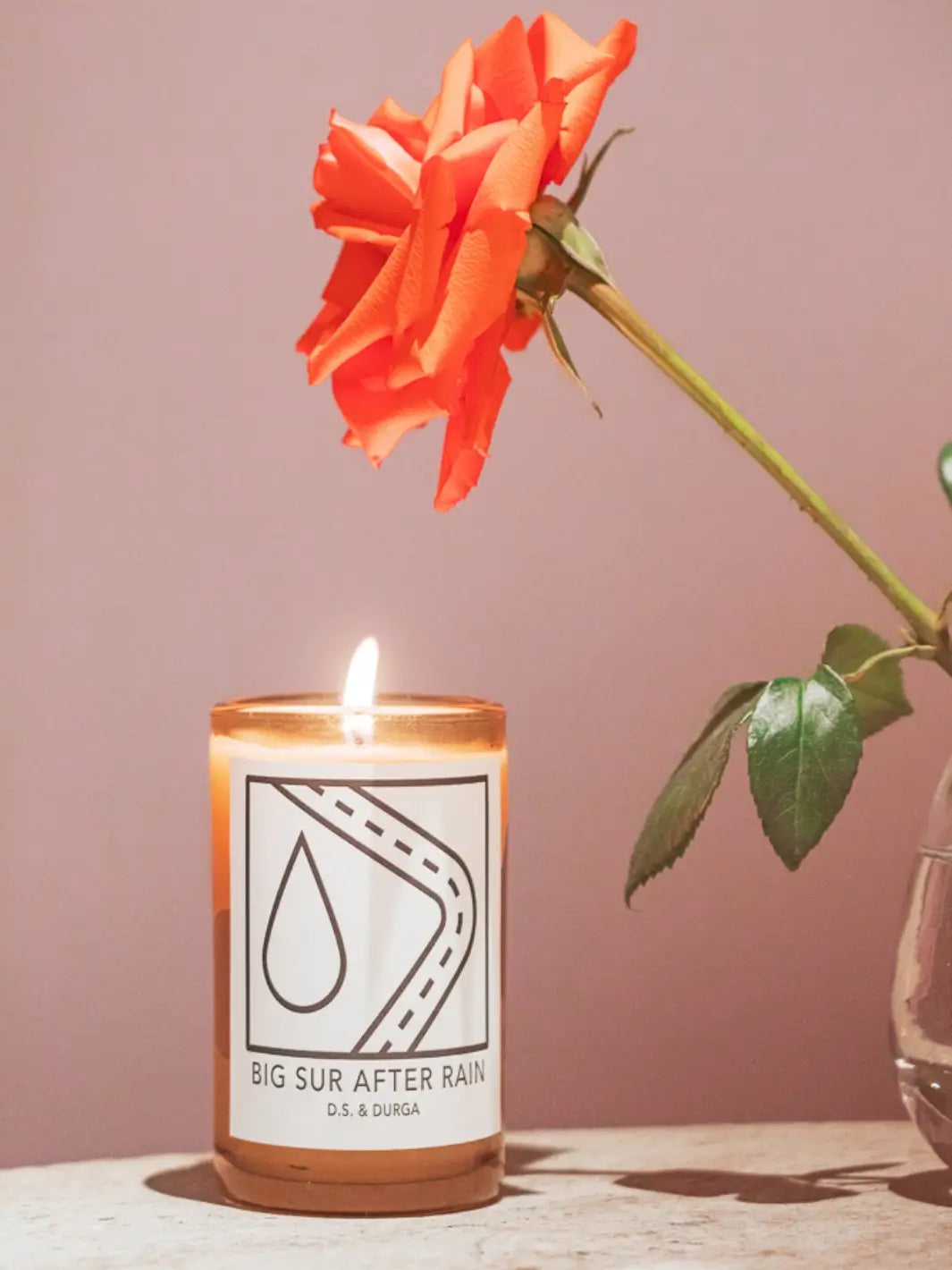 DS&Durga 'Big Sur After Rain' lit candle beside an orange flower in a vase on a pink background | Twentyseven Toronto
