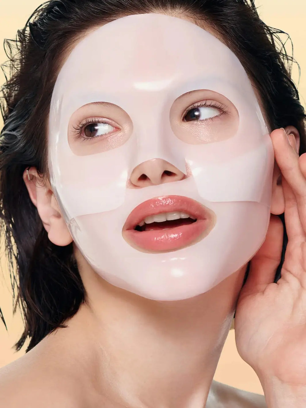 A close-up view shows a woman wearing a thick, white Biodance Bio - Collagen Real Deep Mask sheet on her face against a soft beige background | Twentyseven Toronto