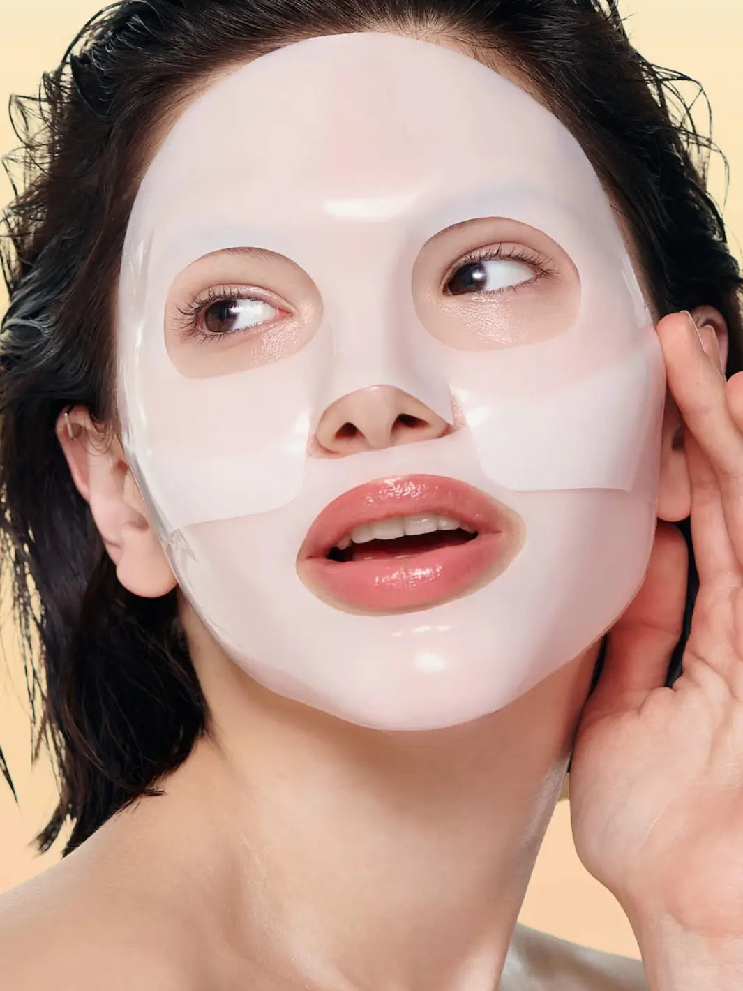 A close-up view shows a woman wearing a thick, white Biodance Bio - Collagen Real Deep Mask sheet on her face against a soft beige background | Twentyseven Toronto