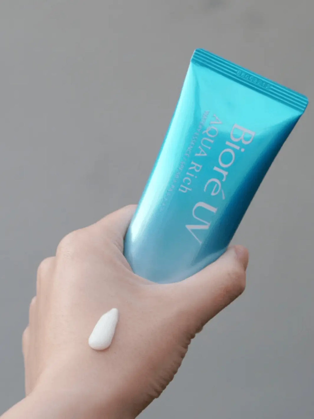 Hand holding Biore UV Aqua Rich Watery Essence SPF50+ PA++++ blue sunscreen tube with product texture swatch | Twentyseven Toronto