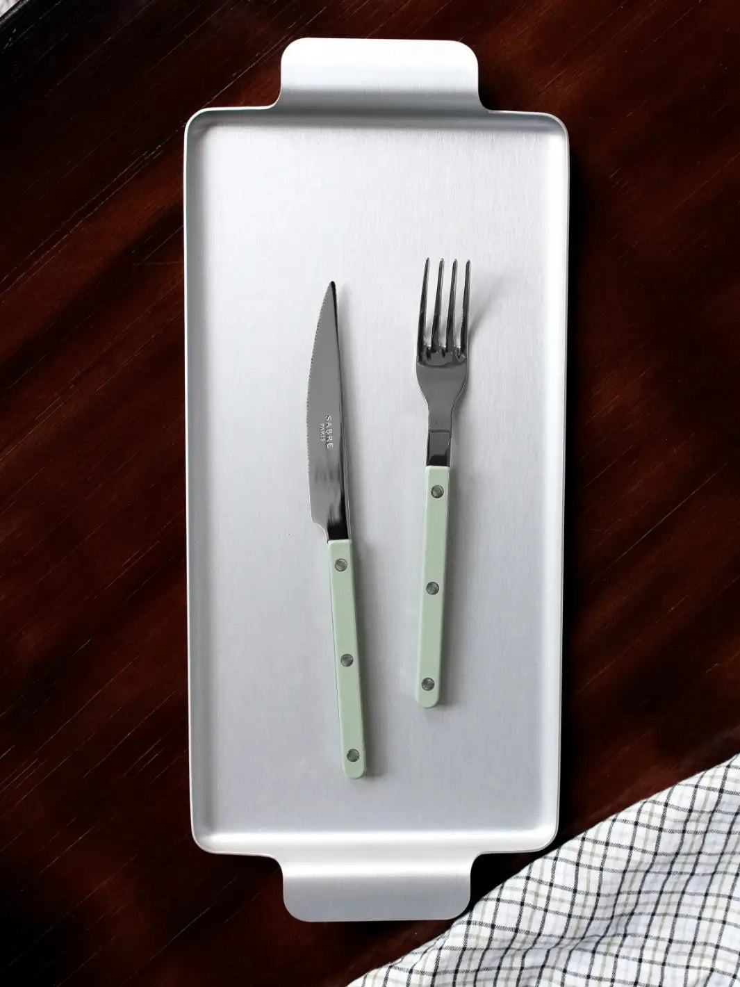 Dinner knife and fork from the Sabre Paris Bistrot Solid Shiny 5 Piece Cutlery Set in Asparagus resting on a rectangular silver serving tray against a dark wood background with a checkered napkin | Twentyseven Toronto