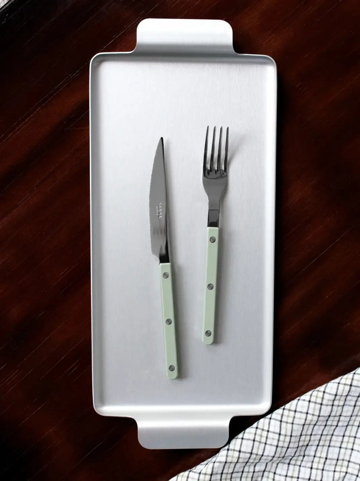 Dinner knife and fork from the Sabre Paris Bistrot Solid Shiny 5 Piece Cutlery Set in Asparagus resting on a rectangular silver serving tray against a dark wood background with a checkered napkin | Twentyseven Toronto