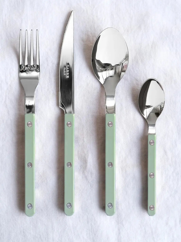 Four pieces from the Sabre Paris Bistrot Solid Shiny 5 Piece Cutlery Set in Asparagus, including a dinner fork, dinner knife, soup spoon, and teaspoon, laid out vertically on a textured white linen surface | Twentyseven Toronto