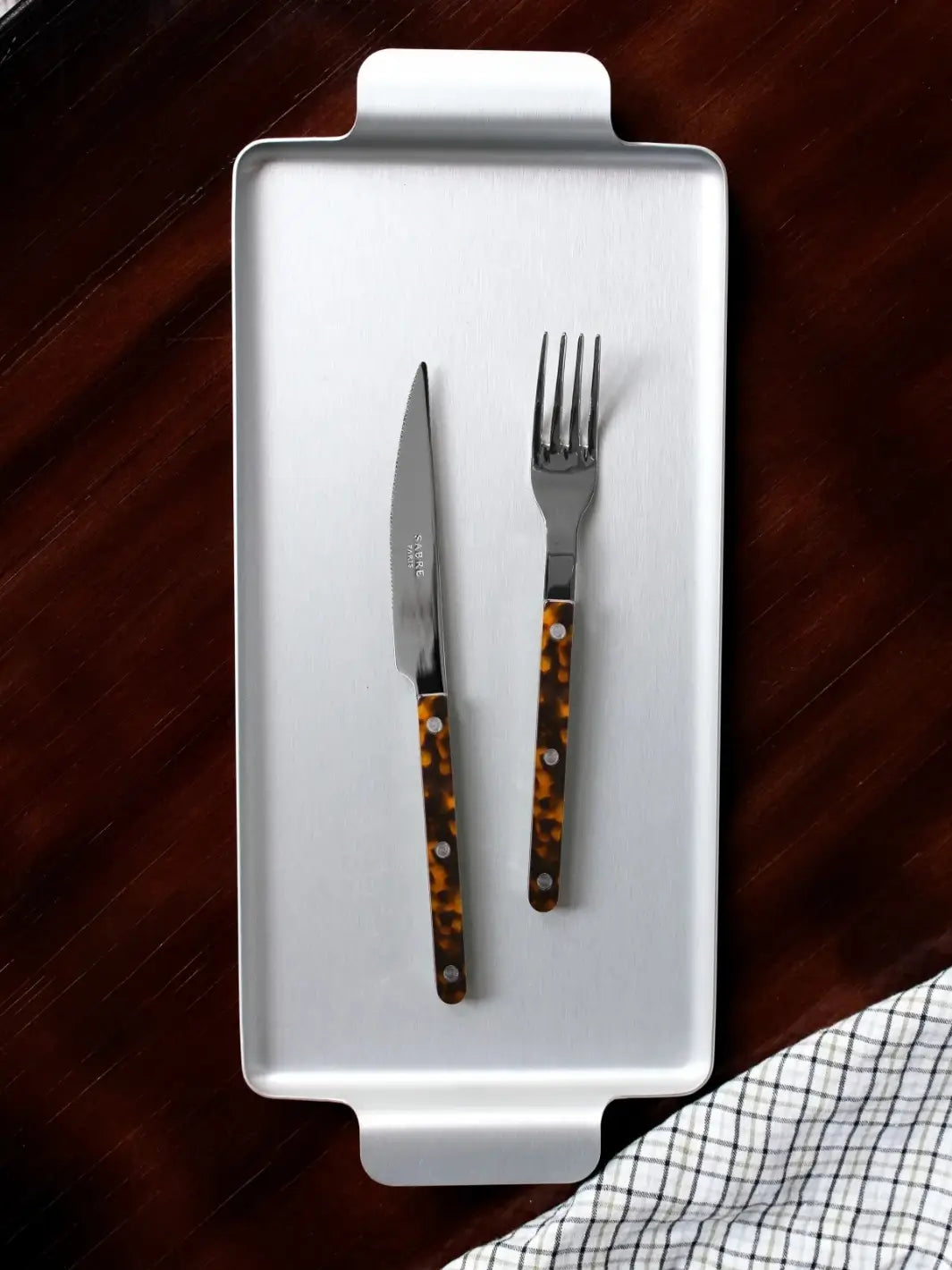 Dinner knife and fork from the Sabre Paris Bistrot Solid Shiny 5 Piece Cutlery Set in Faux Tortoise resting on a rectangular silver serving tray against a dark wood background with a checkered napkin | Twentyseven Toronto