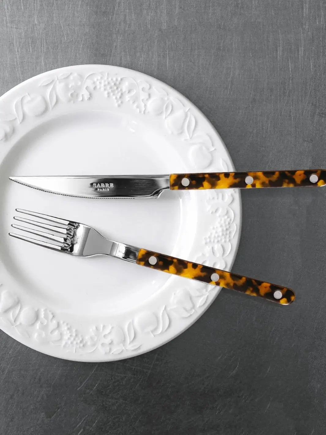 Dinner knife and fork from the Sabre Paris Bistrot Solid Shiny 5 Piece Cutlery Set in Faux Tortoise resting on a white ceramic plate with an embossed border against a dark gray table | Twentyseven Toronto