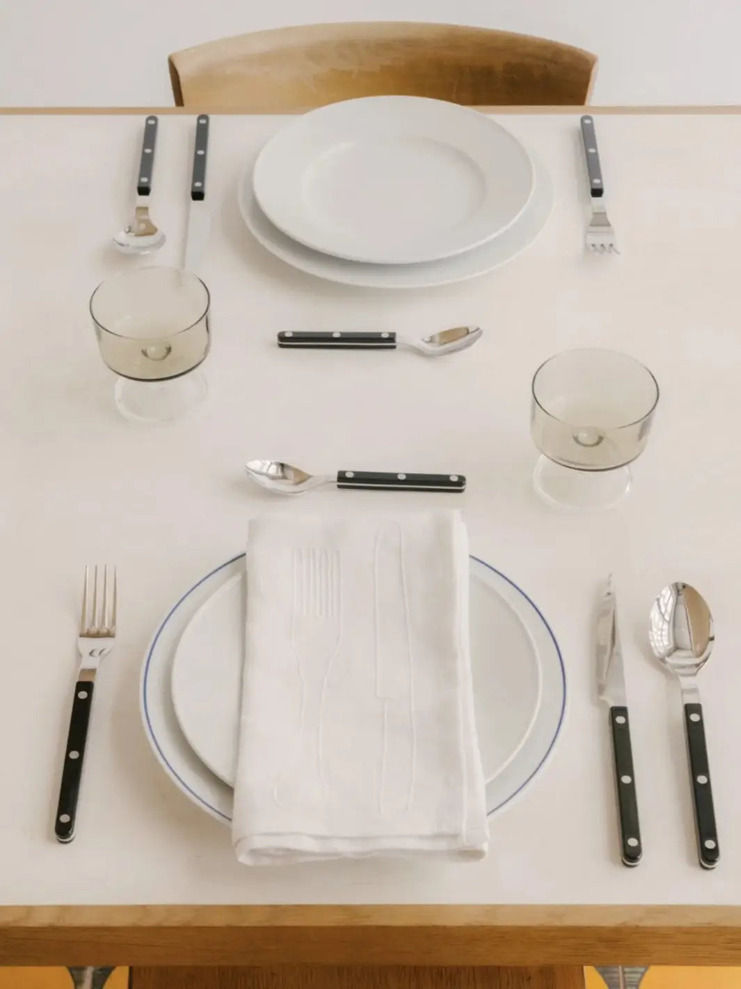 Elegant table setting for two features various pieces from the Sabre Paris Bistrot Solid Shiny 5 Piece Cutlery Set in Black arranged around white plates and smoke-coloured glassware | Twentyseven Toronto
