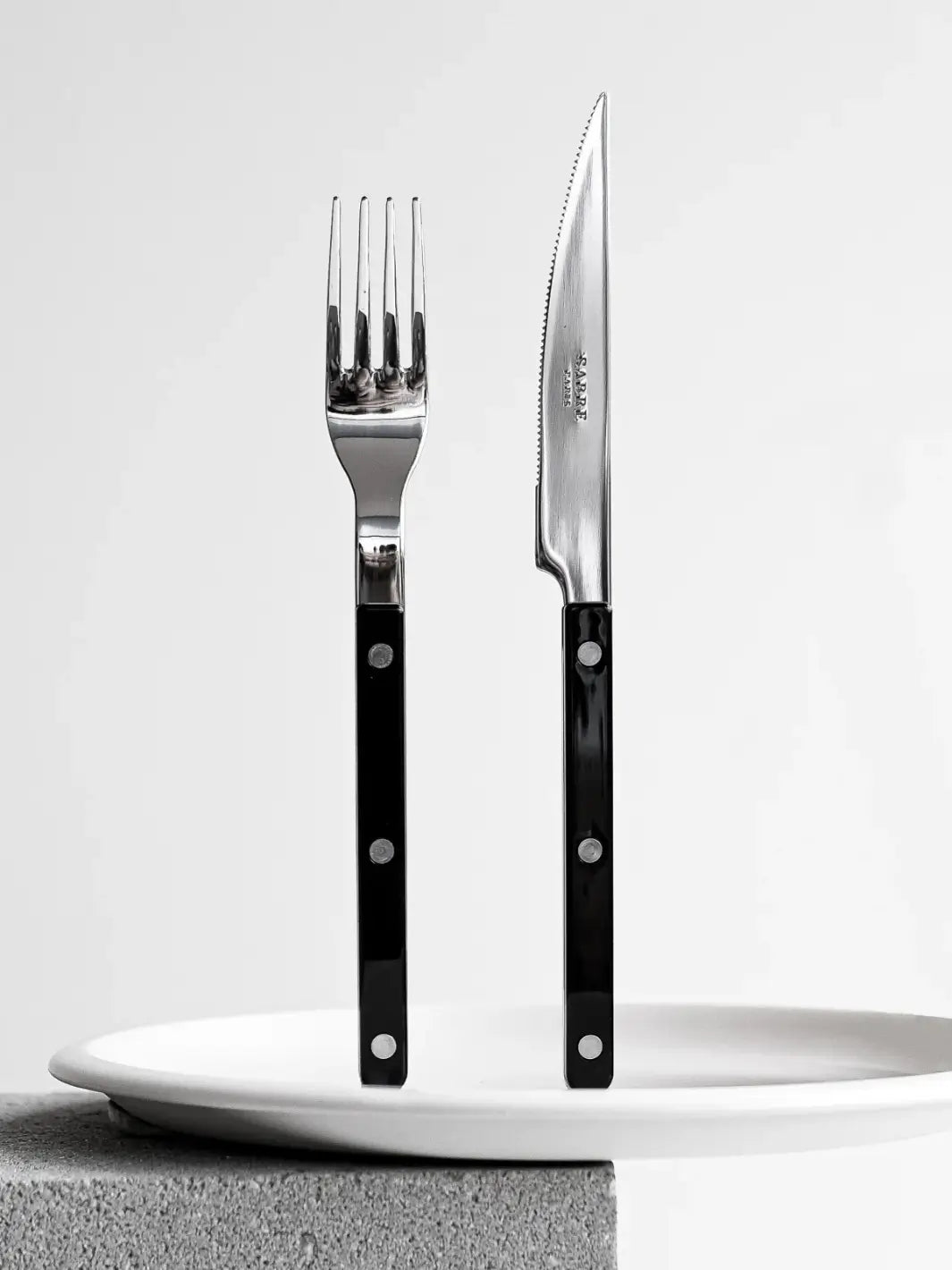 Dinner fork and knife from the Sabre Paris Bistrot Solid Shiny 5 Piece Cutlery Set in Black are shown standing upright behind a stack of white ceramic plates | Twentyseven Toronto