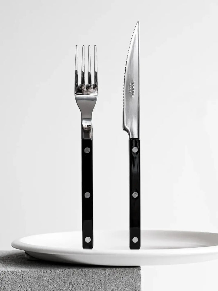 Dinner fork and knife from the Sabre Paris Bistrot Solid Shiny 5 Piece Cutlery Set in Black are shown standing upright behind a stack of white ceramic plates | Twentyseven Toronto