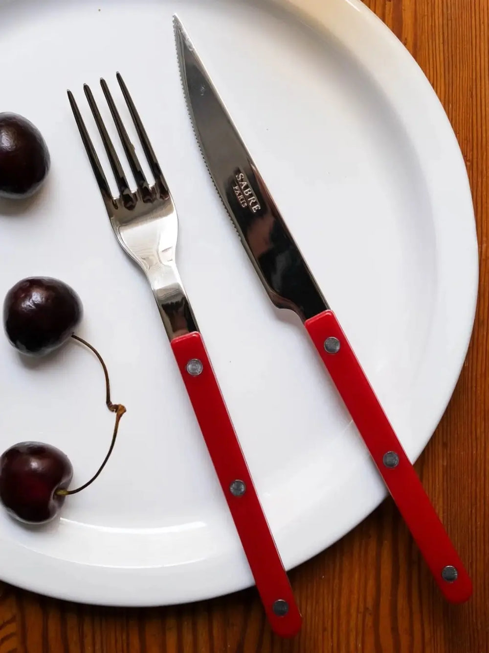 Dinner knife and fork from the Sabre Paris Bistrot Solid Shiny 5 Piece Cutlery Set in Burgundy resting on a white ceramic plate alongside fresh dark cherries | Twentyseven Toronto