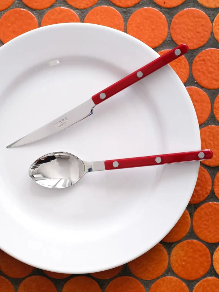 Dinner knife and soup spoon from the Sabre Paris Bistrot Solid Shiny 5 Piece Cutlery Set in Burgundy are displayed on a white plate, against a background of bright orange circular tiles | Twentyseven Toronto