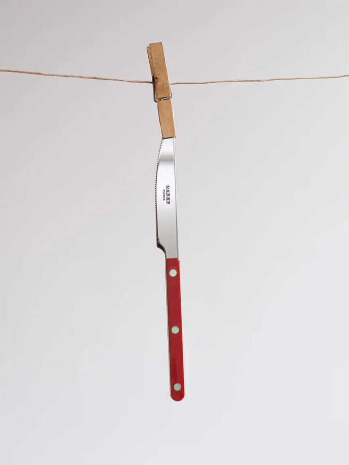 Dinner knife from the Sabre Paris Bistrot Solid Shiny 5 Piece Cutlery Set in Burgundy hanging vertically from a twine clothesline, secured by a wooden clothespin | Twentyseven Toronto