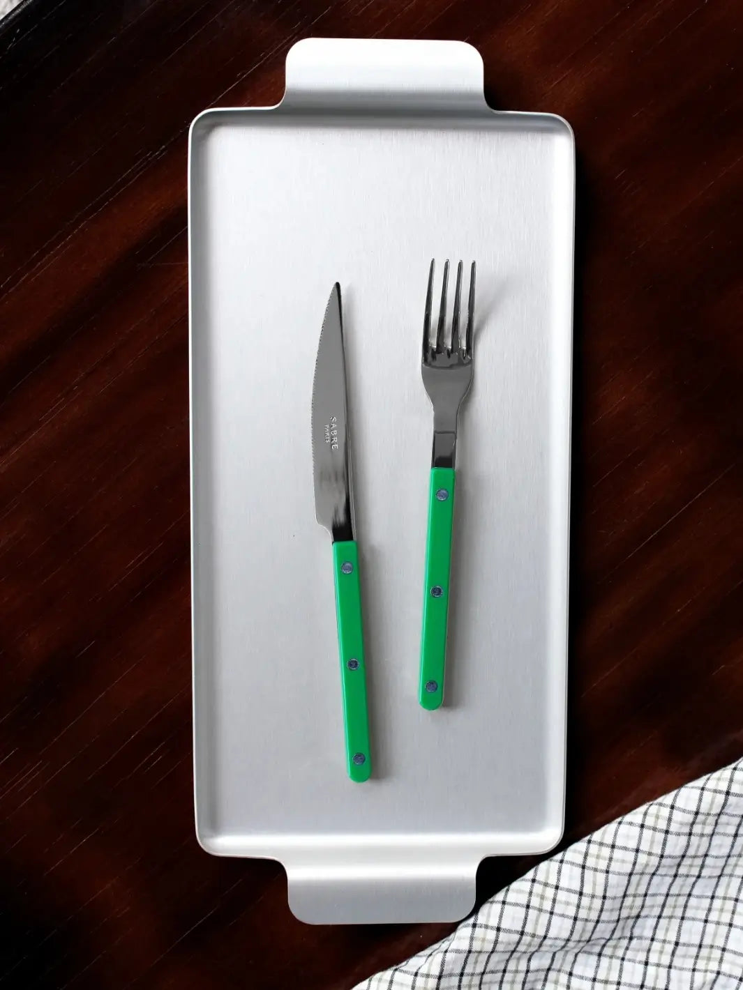 Dinner knife and fork from the Sabre Paris Bistrot Solid Shiny 5 Piece Cutlery Set in Garden Green resting on a rectangular silver serving tray against a dark wood background with a checkered napkin | Twentyseven Toronto
