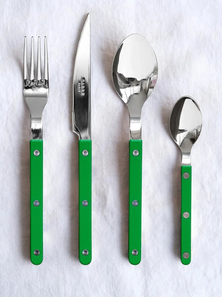 Four pieces from the Sabre Paris Bistrot Solid Shiny 5 Piece Cutlery Set in Garden Green, including a dinner fork, dinner knife, soup spoon, and teaspoon, laid out vertically on a textured white linen surface | Twentyseven Toronto