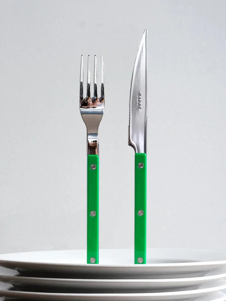 Dinner fork and knife from the Sabre Paris Bistrot Solid Shiny 5 Piece Cutlery Set in Garden Green are shown standing upright behind a stack of white ceramic plates | Twentyseven Toronto