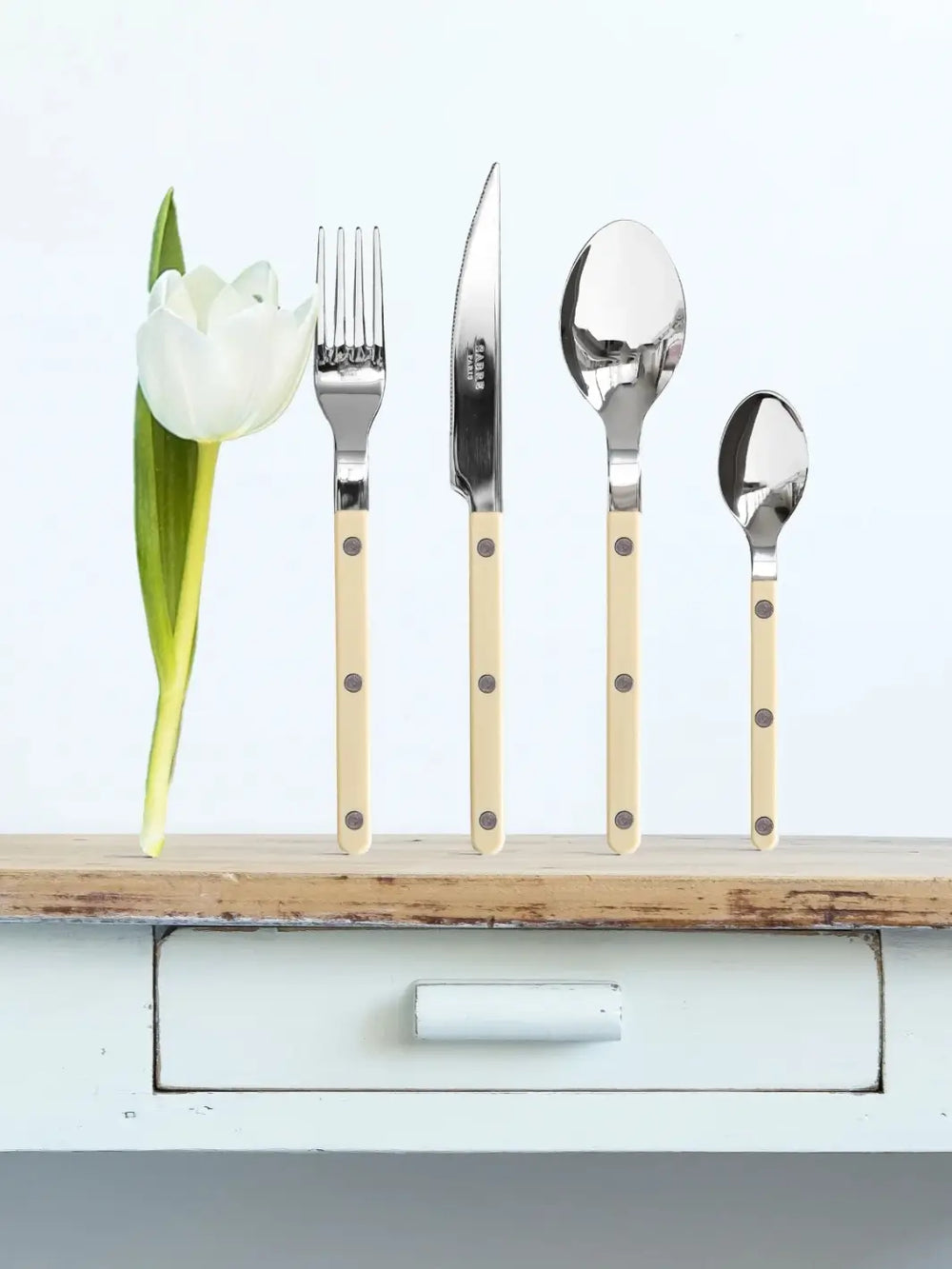 Four cutlery pieces including a soup spoon, a dinner fork, a dinner knife and a teaspoon from the Sabre Paris Bistrot Solid Shiny 5 Piece Cutlery Set in Ivory stands upright on a wooden tabletop next to a single white tulip | Twentyseven Toronto