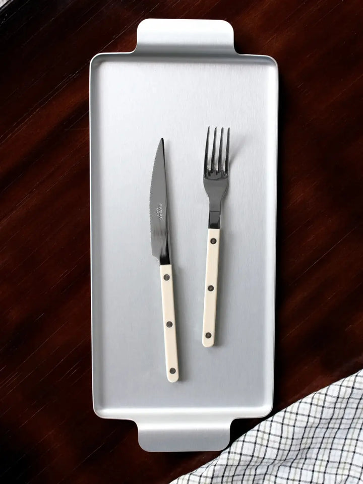 Dinner knife and fork from the Sabre Paris Bistrot Solid Shiny 5 Piece Cutlery Set in Black resting on a rectangular silver serving tray against a dark wood background with a checkered napkin | Twentyseven Toronto