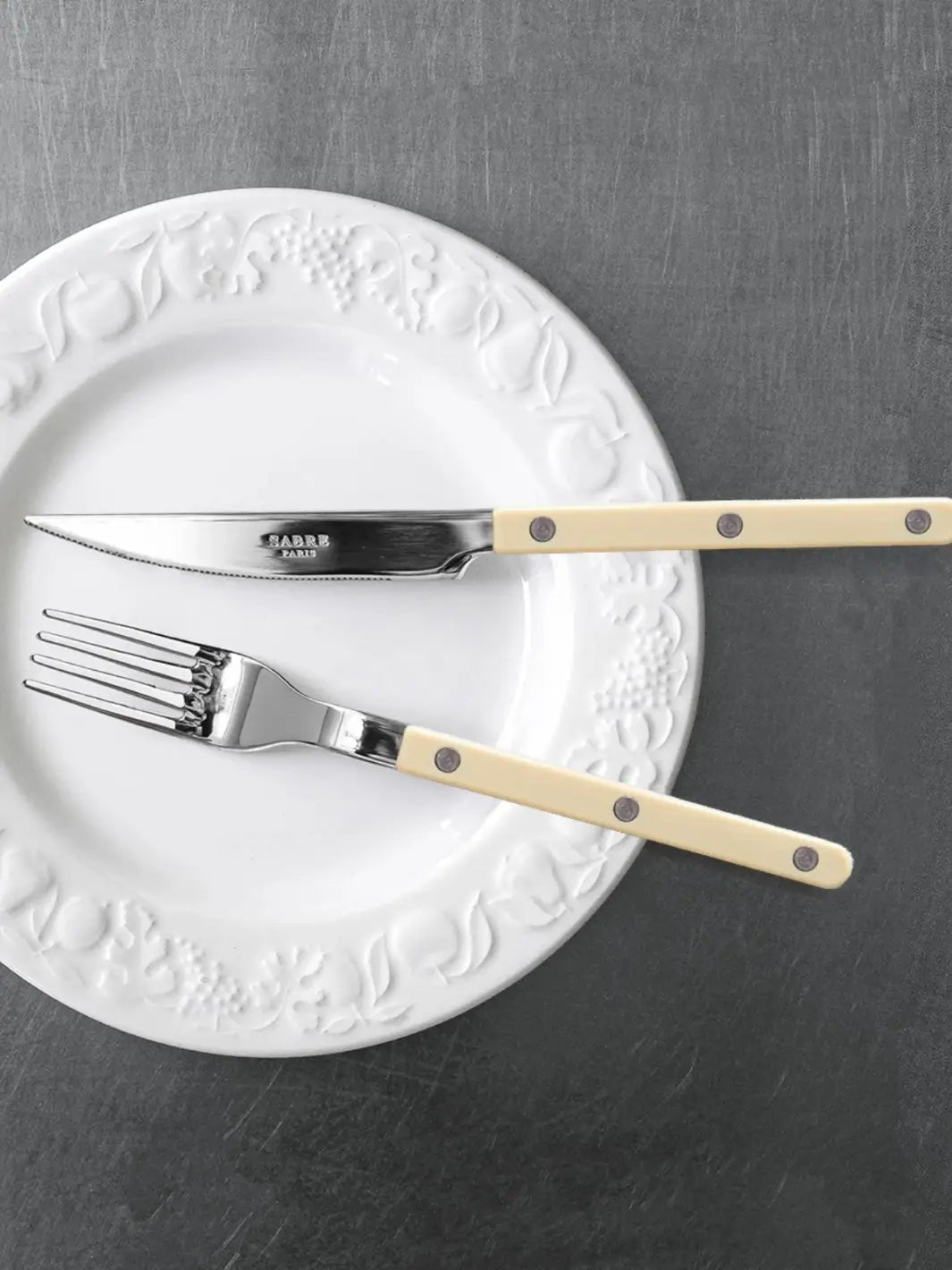 Dinner knife and fork from the Sabre Paris Bistrot Solid Shiny 5 Piece Cutlery Set in Ivory resting on a white ceramic plate with an embossed border against a dark gray table | Twentyseven Toronto