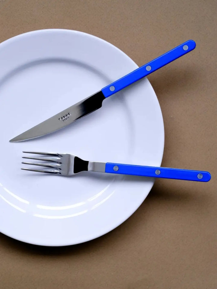 Dinner knife and fork from the Sabre Paris Bistrot Solid Shiny 5 Piece Cutlery Set in Lapis Blue resting on a white plate against a tan background | Twentyseven Toronto
