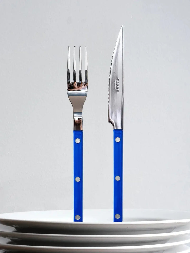 Dinner fork and knife from the Sabre Paris Bistrot Solid Shiny 5 Piece Cutlery Set in Lapis Blue are shown standing upright behind a stack of white ceramic plates | Twentyseven Toronto