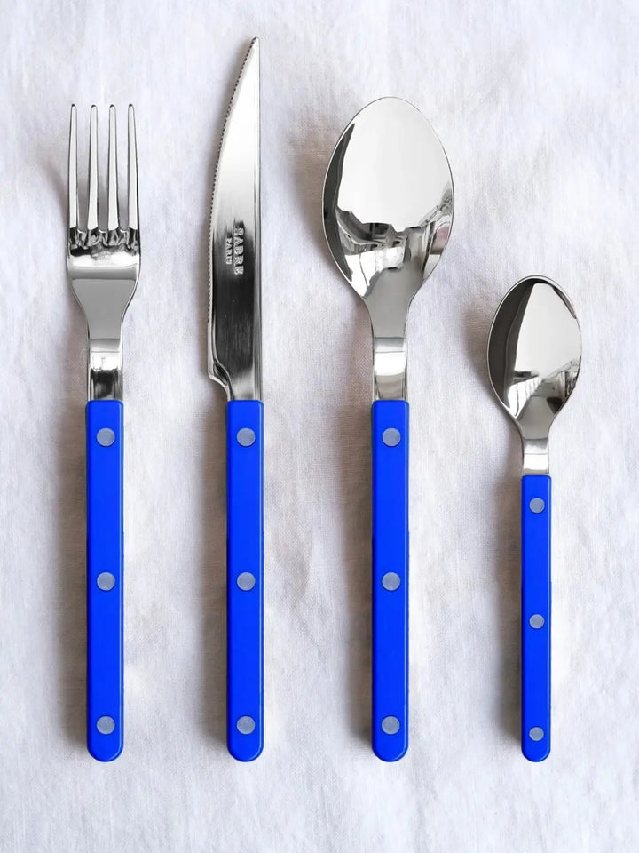 Four pieces from the Sabre Paris Bistrot Solid Shiny 5 Piece Cutlery Set in Lapis Blue, including a dinner fork, dinner knife, soup spoon, and teaspoon, laid out vertically on a textured white linen surface | Twentyseven Toronto