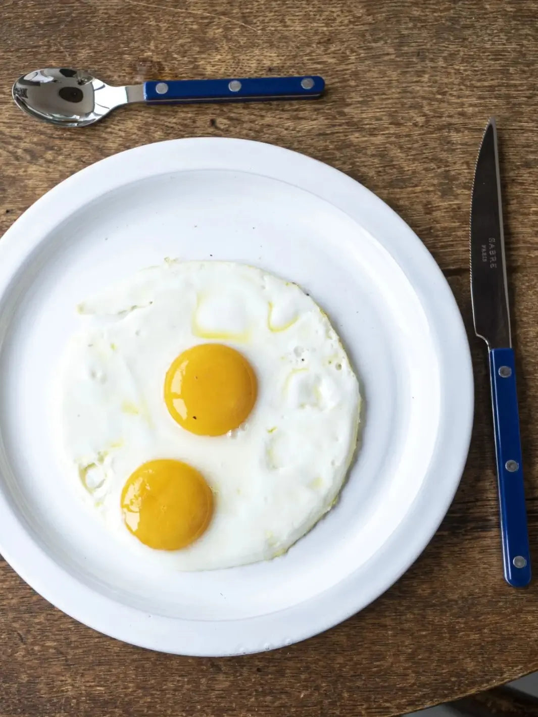 Dinner knife and teaspoon from the Sabre Paris Bistrot Solid Shiny 5 Piece Cutlery Set in Navy Blue arranged around a white plate with two fried eggs on a rustic wooden table | Twentyseven Toronto