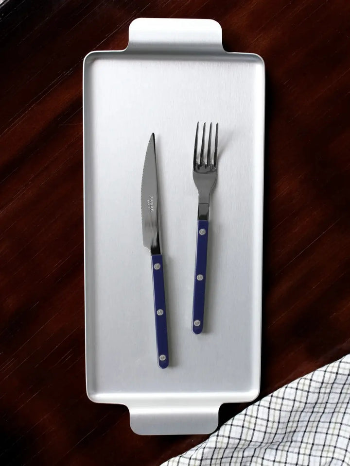 Dinner knife and fork from the Sabre Paris Bistrot Solid Shiny 5 Piece Cutlery Set in Navy Blue resting on a rectangular silver serving tray against a dark wood background with a checkered napkin | Twentyseven Toronto