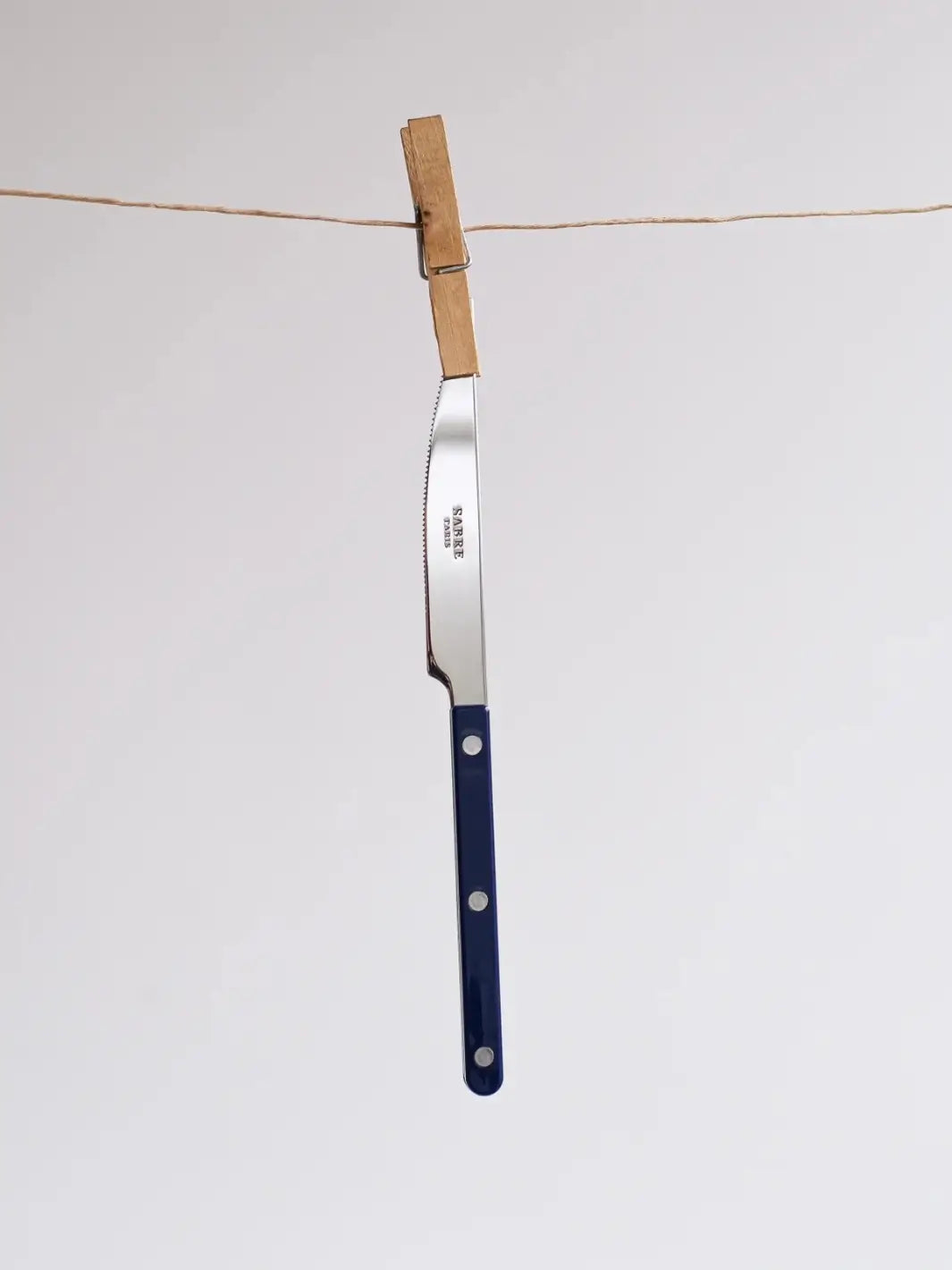 Dinner knife from the Sabre Paris Bistrot Solid Shiny 5 Piece Cutlery Set in Navy Blue hanging vertically from a twine clothesline, secured by a wooden clothespin | Twentyseven Toronto