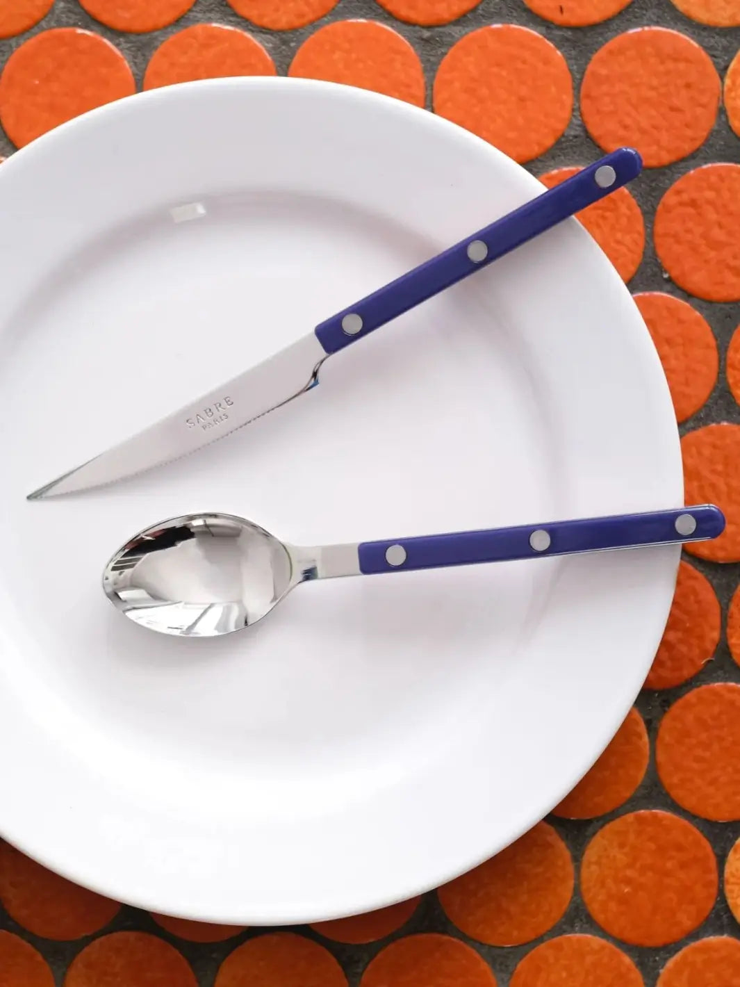 Dinner knife and soup spoon from the Sabre Paris Bistrot Solid Shiny 5 Piece Cutlery Set in Navy Blue are displayed on a white plate, against a background of bright orange circular tiles | Twentyseven Toronto