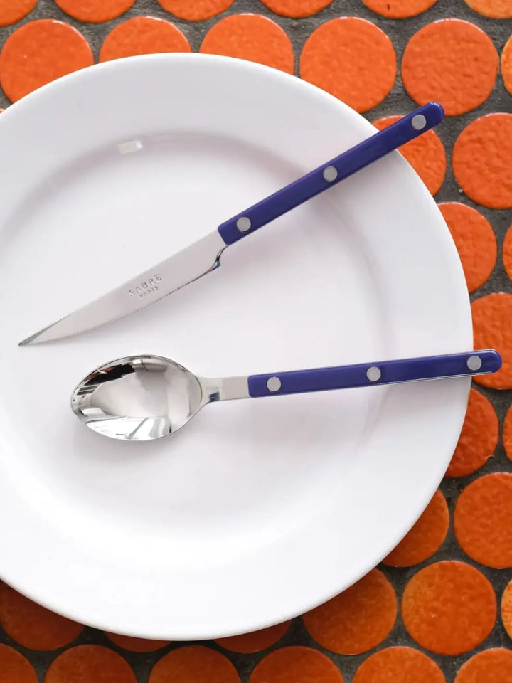 Dinner knife and soup spoon from the Sabre Paris Bistrot Solid Shiny 5 Piece Cutlery Set in Navy Blue are displayed on a white plate, against a background of bright orange circular tiles | Twentyseven Toronto