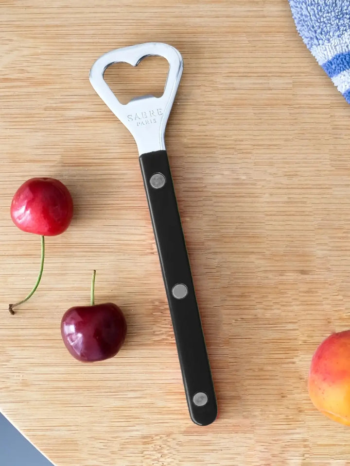 Sabre Paris Shiny Bistrot Solid Bottle Opener in Black shot on a light wood cutting board, surrounded by fresh cherries and a peach | Twentyseven Toronto