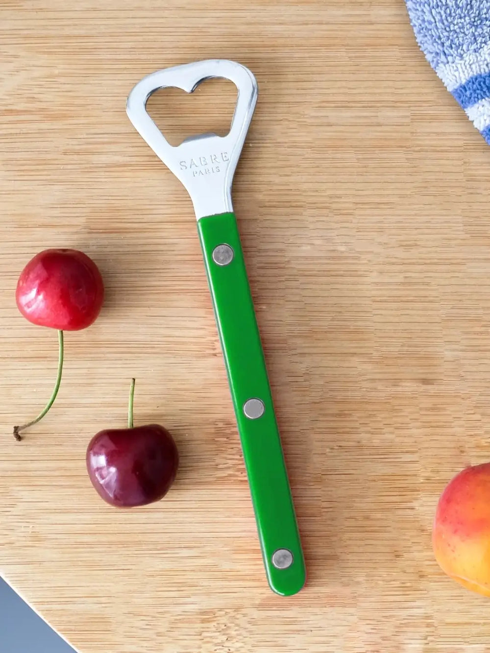 Sabre Paris Shiny Bistrot Solid Bottle Opener in Garden Green shot on a light wood cutting board, surrounded by fresh cherries and a peach | Twentyseven Toronto