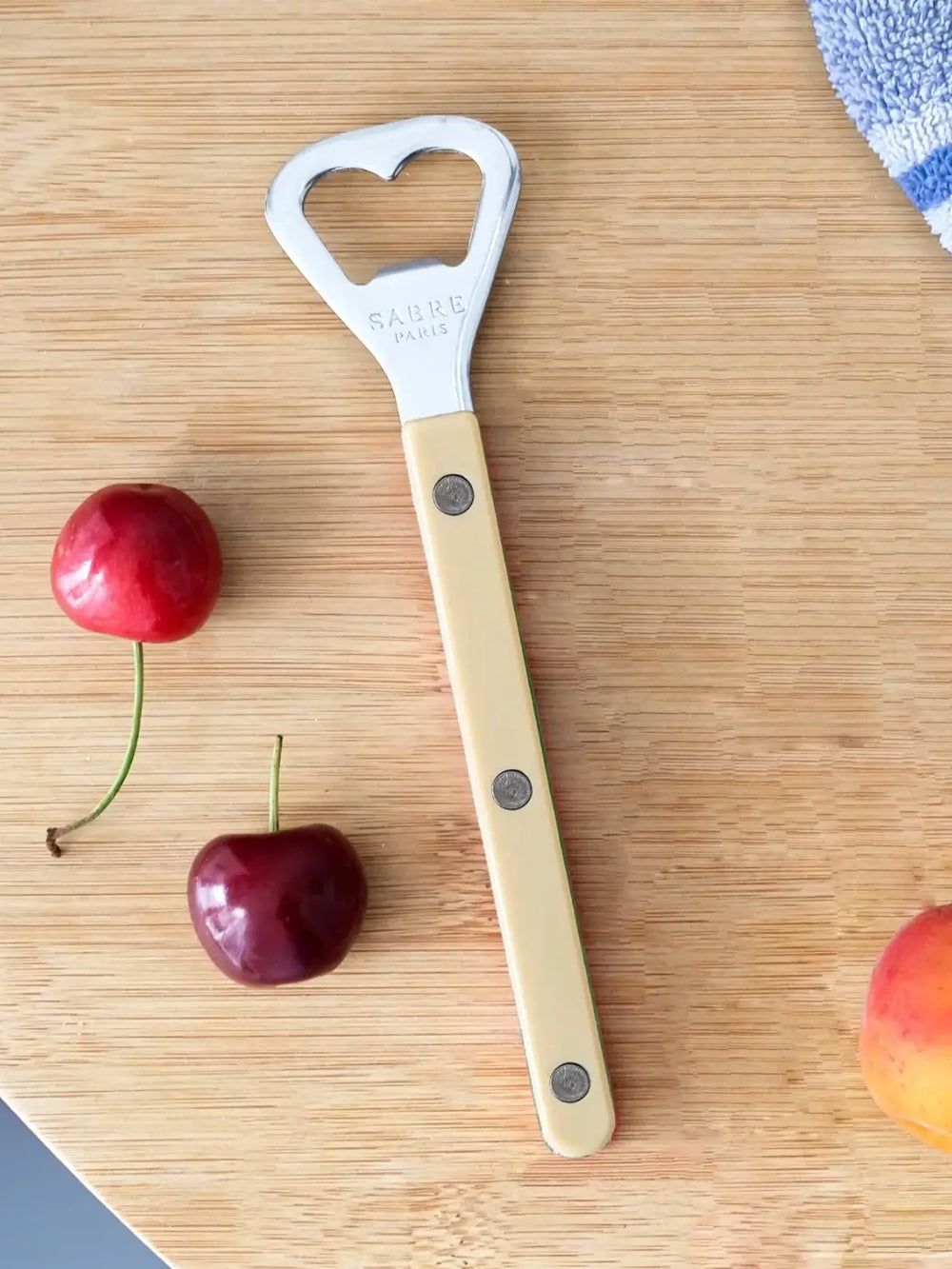 Sabre Paris Shiny Bistrot Solid Bottle Opener in Ivory shot on a light wood cutting board, surrounded by fresh cherries and a peach | Twentyseven Toronto